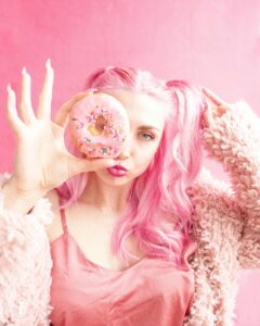 Playful portrait of a woman with pink hair holding a sprinkle donut against a pink background.